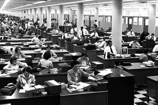 A 1950's photo from a large open office - employees work at rows of wooden desks set up close together that stretch on into the background seemingly infitinely.