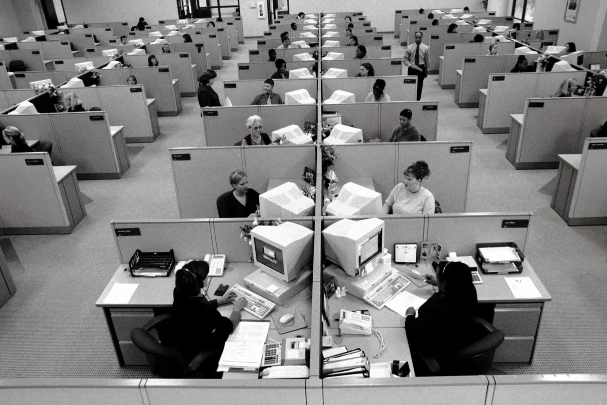 A photo from the 1900s of an office set up with many cubicles. Employees work at their desks on large white computers.